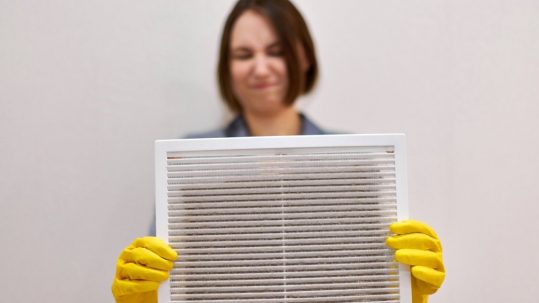 A short-haired woman holding a dirty ventilation grill. She wears yellow gloves and has a disgusted look on her face.