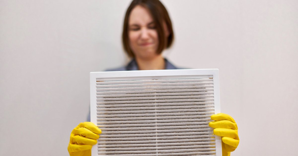 A short-haired woman holding a dirty ventilation grill. She wears yellow gloves and has a disgusted look on her face.