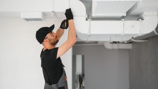 A man fixing an air duct system installed in the ceiling. He's wearing black gloves, a hat, and safety glasses.