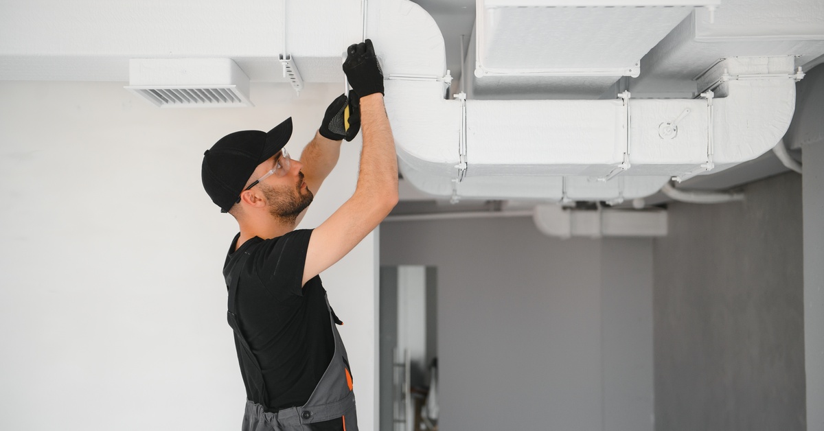 A man fixing an air duct system installed in the ceiling. He's wearing black gloves, a hat, and safety glasses.