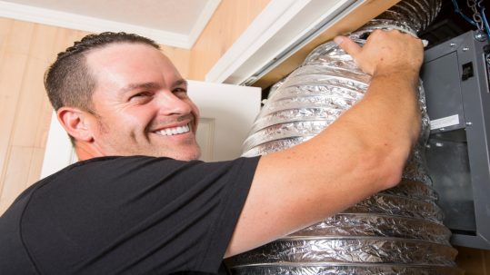 A man in a black shirt smiles while working on a large, flexible air duct connected to an indoor HVAC unit.