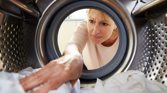 A middle-aged blond woman in a pink shirt reaching into a clothes dryer to grab clean white clothing.