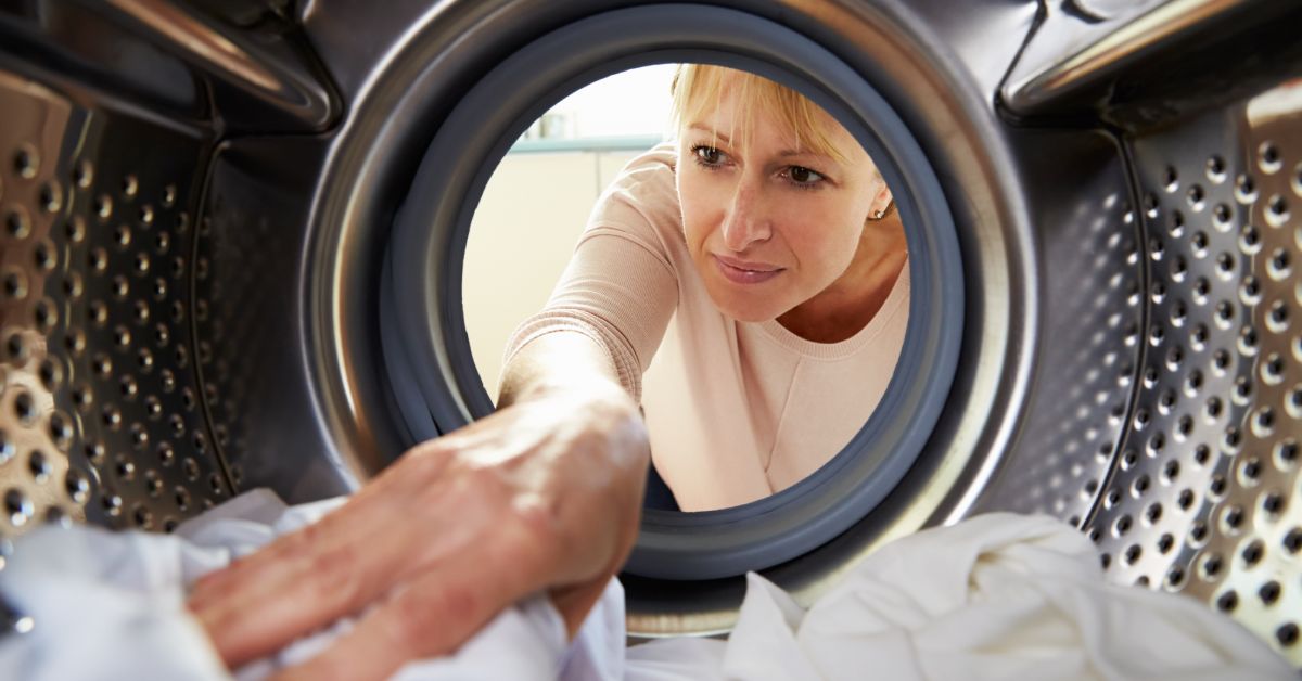 A middle-aged blond woman in a pink shirt reaching into a clothes dryer to grab clean white clothing.