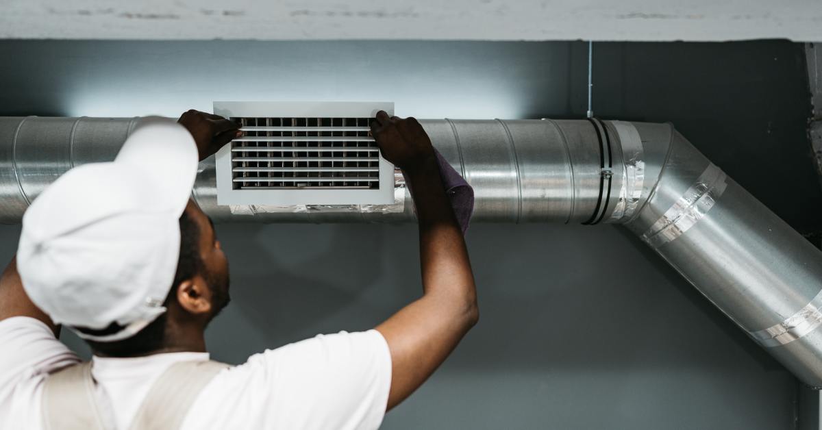 A male HVAC technician installing a white air vent grille on a ductwork system in a commercial building.